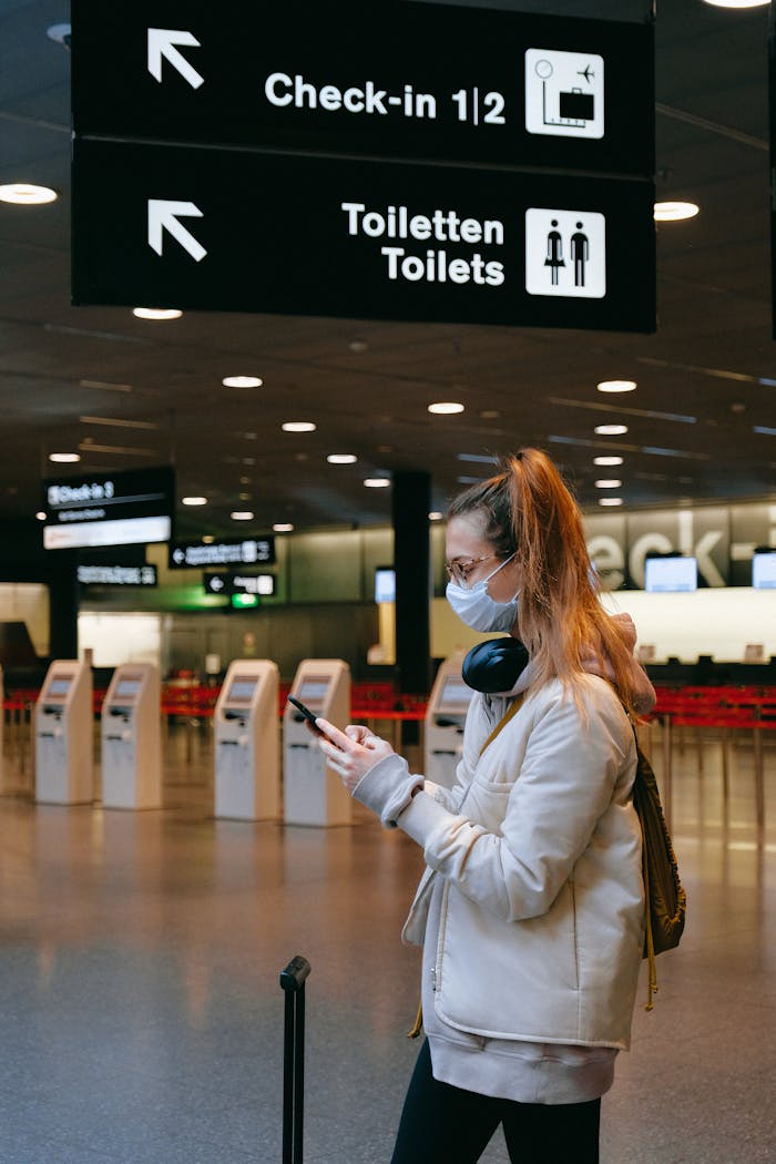 Home Woman wearing a mask, texting in airport terminal with signage in background.